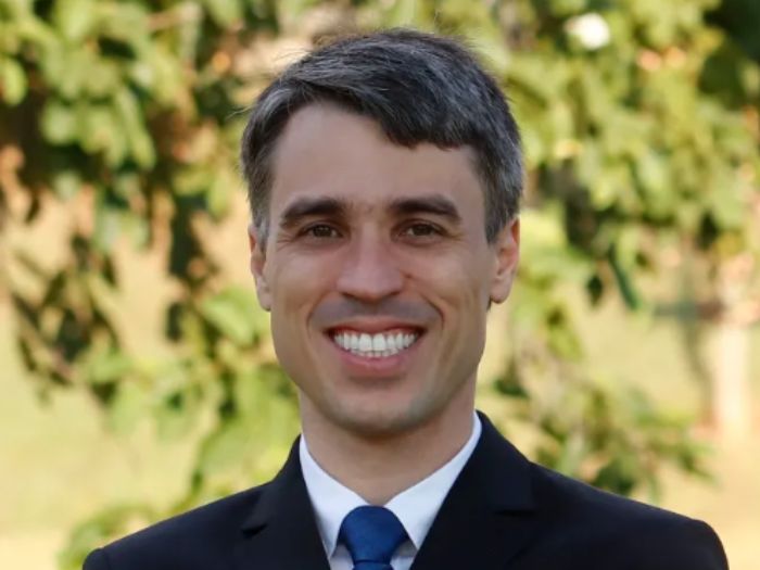 Retrato profissional do Dr. Pedro Rafael de Moura Meireles, sorrindo, vestindo terno escuro e gravata azul, fundo natural com folhagem verde desfocada, iluminação natural suave, fotografia realista